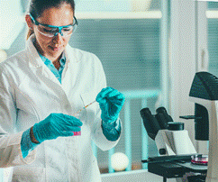 Foto de investigadora en un laboratorio frente a un microscopio, tiene bata blanca, lentes de protección, guantes.