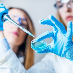 Foto de un par de jóvenes en un laboratorio haciendo un experimento, ambas con guantes de látex y anteojos protectores.