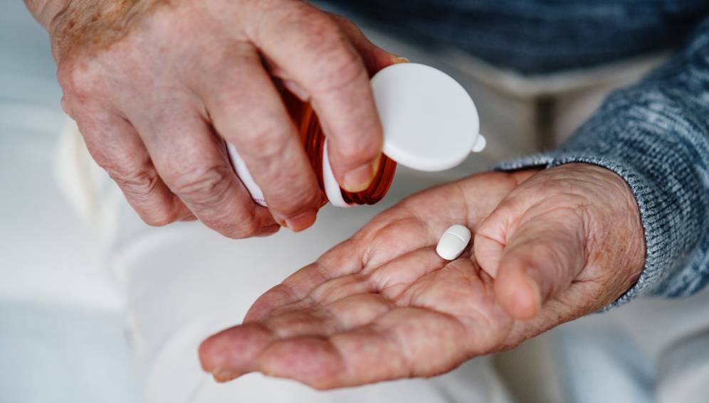 Foto de dos manos de adulto mayor, en una mano el frasco de pastillas y en la otra una pastilla blanca.