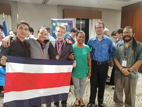 Seis personas posan sonrientes dentro una sala, tres estudiantes sostienen la bandera de Costa Rica, dos portan sus medallas al cuello. 