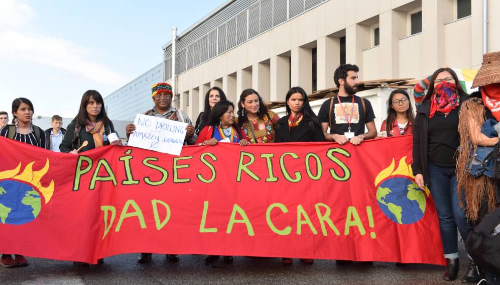En la foto se ve a un grupo de personas de pie sosteniendo una gran manta que dice "Países ricos dad la cara".