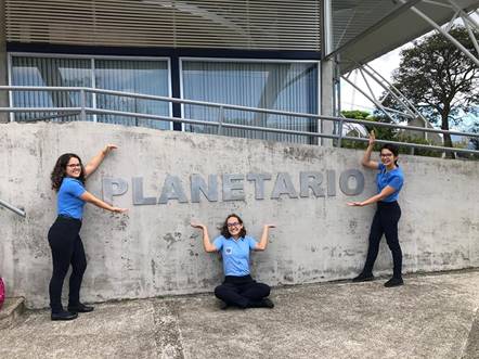 Tres estudiantes del Colegio Científico, posan en el exterior junto al rótulo del Planetario, dos de pie y una sentada, todas sonríen.