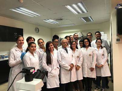 El Dr. Chinchilla posa de pie junto a otras 16 personas, todos con gabachas blancas, que son el personal académico y estudiantes del Laboratorio de Ciencia Básica UCIMED