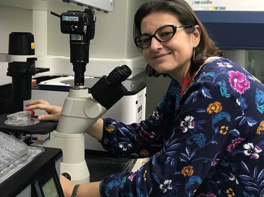 Foto de la Dra. Corrales posa sentada frente a un microscopio y sonríe.